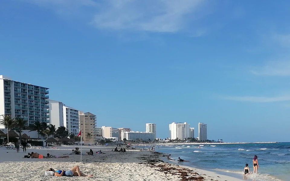 Playa Delfines: arena blanca con bañistas, el mar turquesa a la derecha y la zona hotelera de Cancún alineada al fondo