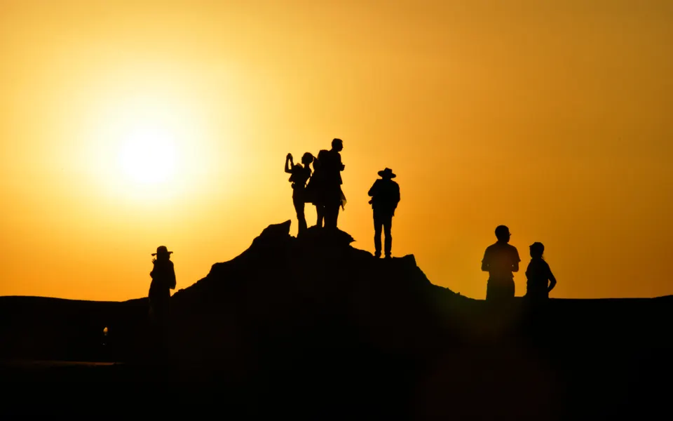 Siete personas en silueta sobre una loma de arena contra el sol poniente, de estaturas muy distintas: unas altas juntas arriba, otras más bajas a los lados