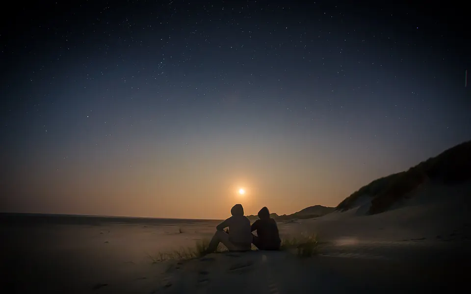 Dos personas sentadas juntas en la arena, en silueta, mirando la última luz sobre el mar bajo un cielo estrellado